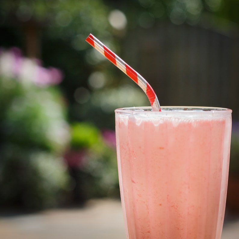 May include: A pink smoothie in a glass with a red and white striped straw.
