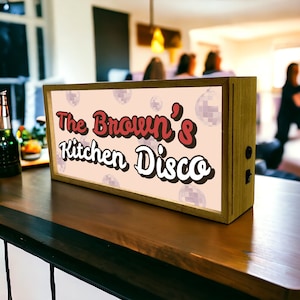 May include: A wooden sign with a pink background and white disco ball designs. The sign says "The Brown's Kitchen Disco" in red and white lettering.