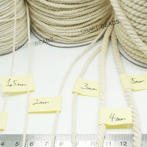 Pu&ograve; includere: Cinque bobine di corda di cotone naturale in diverse dimensioni, da 1 mm a 5 mm, esposte su una superficie bianca con un righello di riferimento. La corda &egrave; non sbiancata e ha un aspetto naturale e rustico.