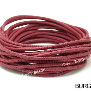 May include: A close-up of a spool of burgundy waxed cotton cord. The cord is tightly wound and has a textured surface. The text "CRAFT BEADS" is visible on the spool.