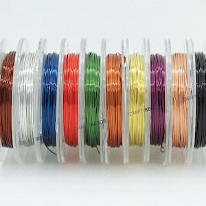 Op de afbeelding: Een verzameling kleurrijke ambachtelijke draadspoelen in verschillende kleuren, waaronder bruin, zilver, blauw, rood, groen, oranje, geel, paars, koper en zwart. Elke spoel is netjes opgerold en wordt weergegeven op een doorzichtige plastic houder. De tekst "CRAFT BEADS" is zichtbaar.