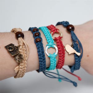 May include: Five braided bracelets with different colored cords and charms. The bracelets include a camera charm, a bird charm, and two silver rings. The bracelets are on a person's wrist.