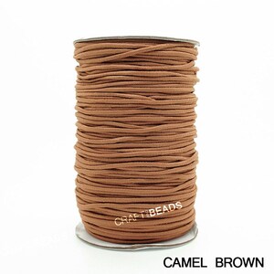 May include: A spool of camel brown elastic cord, wound tightly on a white cardboard spool. The cord is smooth and has a matte finish. The text "CRAFT BEADS" is printed on the spool in gold lettering. The text "CAMEL BROWN" is printed below the spool in black lettering.