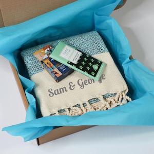May include: A gift box containing a patterned towel, a bar of dark chocolate, and a small box of chocolates. The towel is cream and teal with the text "Sam & George". The box is lined with blue tissue paper.
