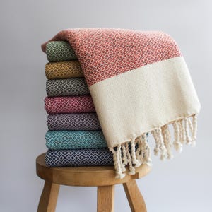 May include: A stack of colourful, diamond-patterned Turkish towels in various colours, including red, green, yellow, grey, and blue. One towel is draped over the stack, featuring a cream-coloured border and fringe.