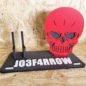 May include: A black wooden stand with two slots for holding items. The stand has a red skull cutout on top and the text "_J03F4RROW_" on the front.