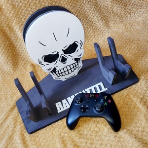 May include: Black skull-shaped video game controller stand with the text "RAVEN TIII" on the front. A black Xbox controller is resting on the stand.