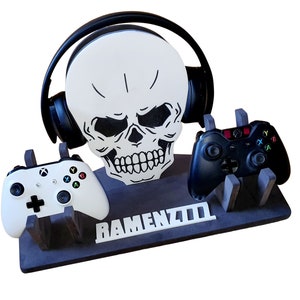 May include: A black and white skull-shaped gaming console stand with two controllers and a headset. The stand has the word "RAMENZ700" written on it.