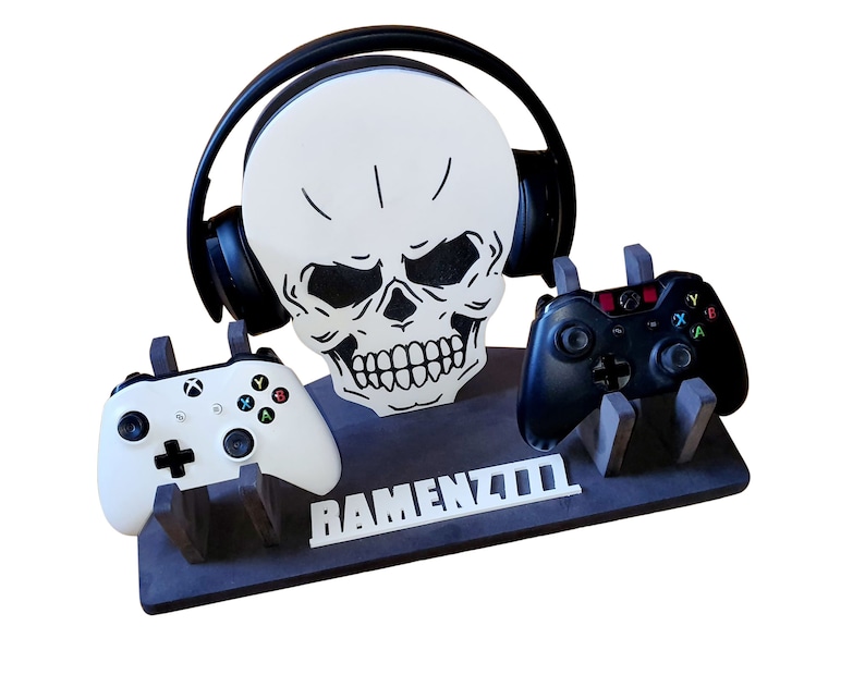 May include: A black and white skull-shaped gaming console stand with two controllers and a headset. The stand has the word "RAMENZ700" written on it.
