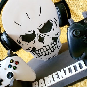 May include: A black and white skull-shaped stand with a white inscription that reads "RAMENZ400". The stand holds two black and white video game controllers and a pair of black headphones.