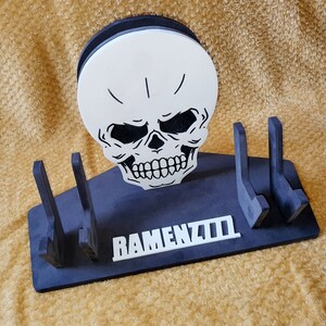 May include: Black wooden stand with three slots for holding items. The stand features a white skull with black eyes and a white inscription that reads "RAMENZILLA".