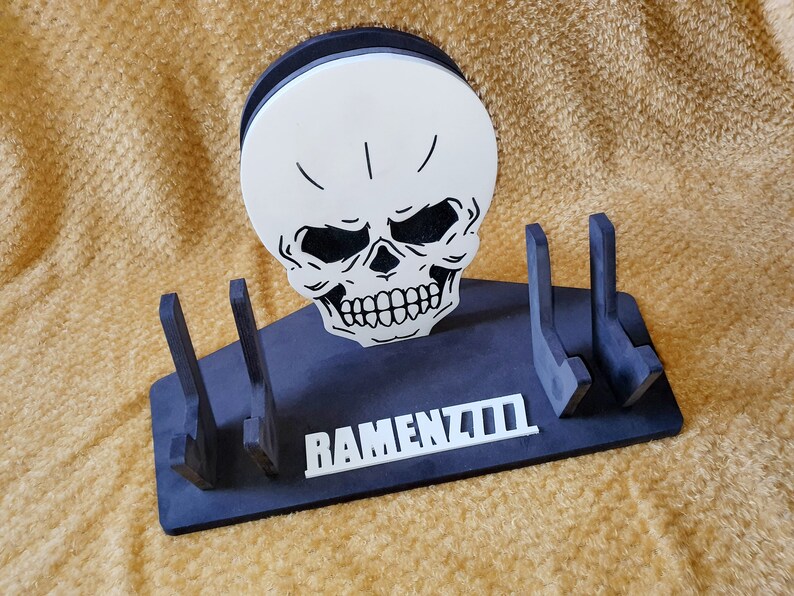 May include: Black wooden stand with three slots for holding items. The stand features a white skull with black eyes and a white inscription that reads "RAMENZILLA".