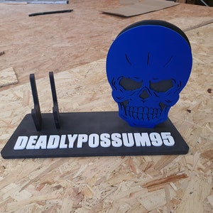 May include: A black and white skull with a blue face and the text "DEADLYPOSSUM95" on a black base. The skull is a stand for two black knife holders.