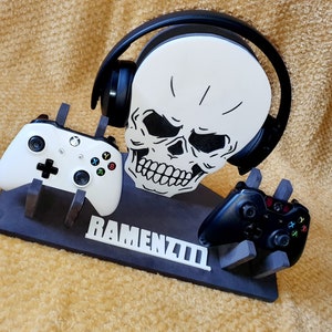 May include: A black and white skull-shaped gaming headset stand with two controllers, one white and one black, and the text "RAMENZIII" on the base.