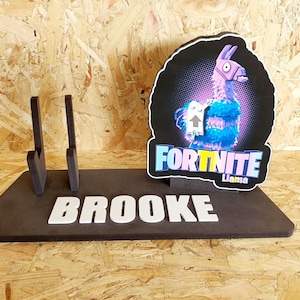 May include: A black wooden stand with the name "BROOKE" in white letters. The stand has two slots for controllers and a Fortnite Llama graphic with the text "Fortnite Llama".