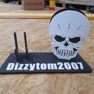 May include: A black and white skull with a toothy grin is mounted on a black stand with the text "Dizzytom2007" in white letters. The stand has two vertical slots on the left side.