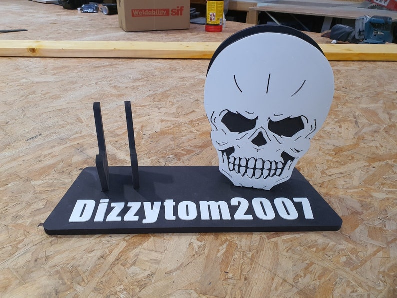 May include: A black and white skull with a toothy grin is mounted on a black stand with the text "Dizzytom2007" in white letters. The stand has two vertical slots on the left side.