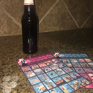 May include: A dark glass bottle with a white label stands next to two "Bachelorette Bingo" game cards. The cards feature images and text, with clear glass gems as markers. The game cards are pink and blue.
