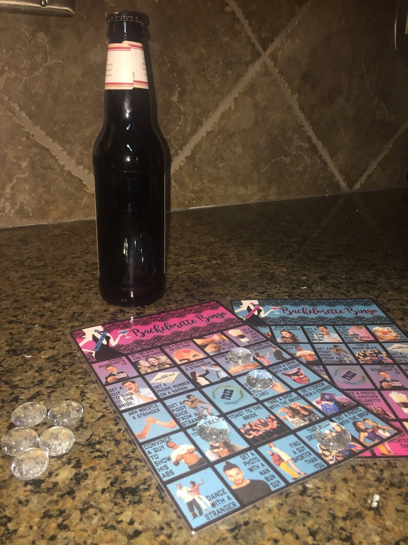 May include: A dark glass bottle with a white label stands next to two "Bachelorette Bingo" game cards. The cards feature images and text, with clear glass gems as markers. The game cards are pink and blue.