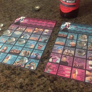 May include: Two "Bachelorette Bingo" game cards with clear glass markers. The cards have a blue and pink color scheme with various images and text. A bottle of beer is visible in the background.
