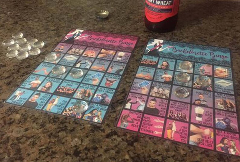 May include: Two "Bachelorette Bingo" game cards with clear glass markers. The cards have a blue and pink color scheme with various images and text. A bottle of beer is visible in the background.