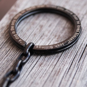 Pode incluir: Porta-chaves de metal preto com um design circular. O porta-chaves apresenta a cita&ccedil;&atilde;o "Um c&atilde;o &eacute; a &uacute;nica coisa na terra que te ama mais do que a si mesmo" de Josh Billings gravada.