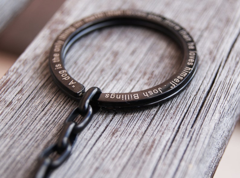 Pode incluir: Porta-chaves de metal preto com um design circular. O porta-chaves apresenta a cita&ccedil;&atilde;o "Um c&atilde;o &eacute; a &uacute;nica coisa na terra que te ama mais do que a si mesmo" de Josh Billings gravada.