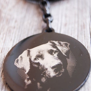 Pode incluir: Um porta-chaves de metal preto com um pendente circular com uma imagem gravada do rosto de um c&atilde;o. O porta-chaves tem um anel e um elo de corrente. O rosto do c&atilde;o &eacute; detalhado com grava&ccedil;&atilde;o clara.