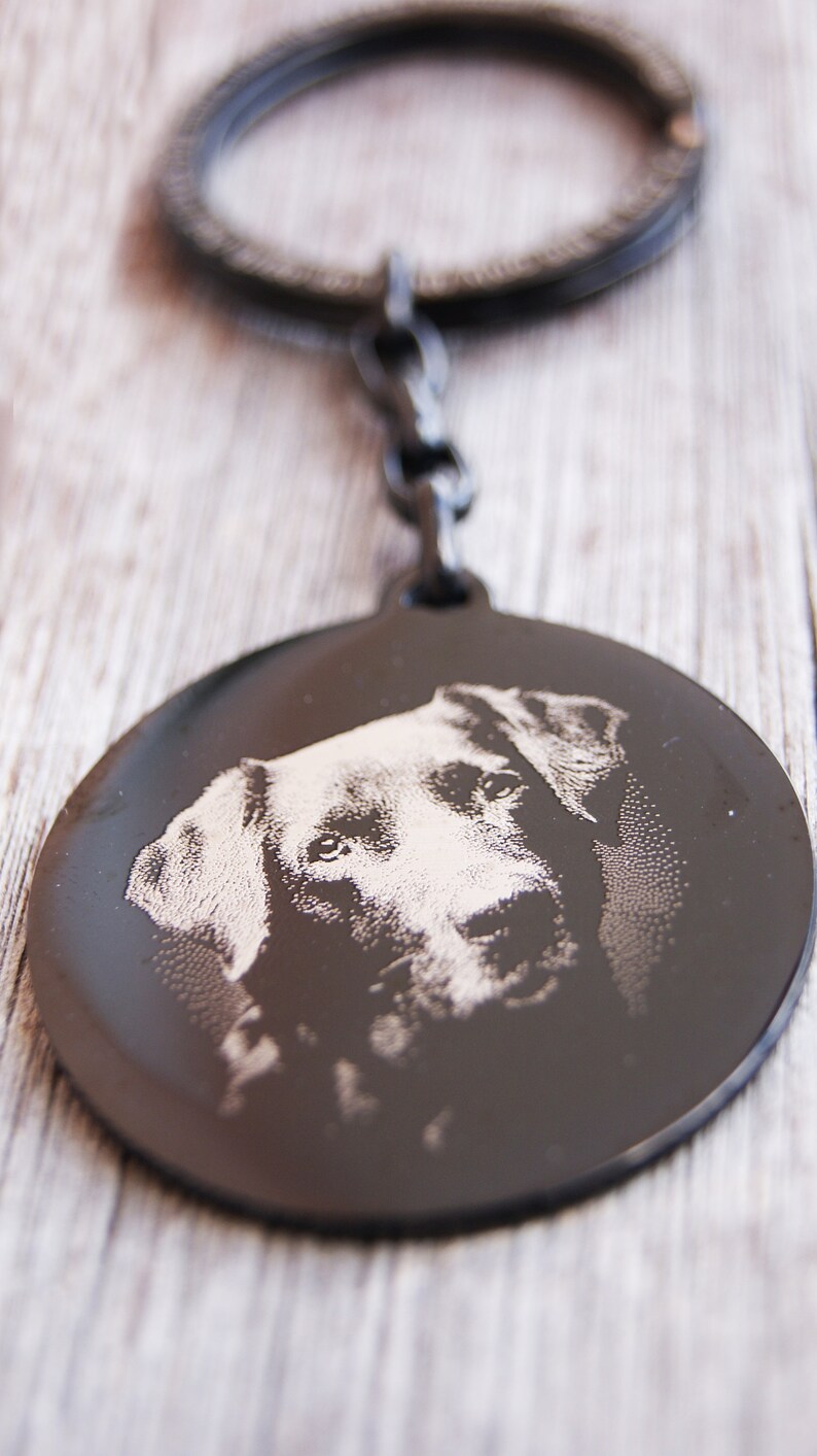 Pode incluir: Um porta-chaves de metal preto com um pendente circular com uma imagem gravada do rosto de um c&atilde;o. O porta-chaves tem um anel e um elo de corrente. O rosto do c&atilde;o &eacute; detalhado com grava&ccedil;&atilde;o clara.