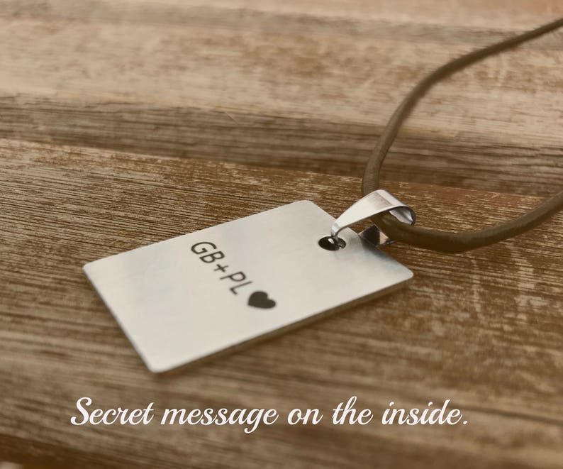Pu&ograve; includere: Una collana d'argento con un ciondolo rettangolare che dice "GB+PL" con un piccolo simbolo di cuore. La collana &egrave; su una superficie di legno marrone.