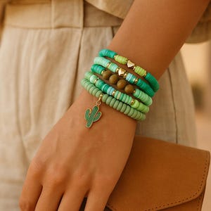 Könnte beinhalten: Mehrere grüne Perlenarmbänder in verschiedenen Farbtönen, verziert mit goldfarbenen Perlen und einem Kaktus-Anhänger. Die Armbänder werden am Handgelenk getragen, im Hintergrund eine braune Clutch. Ein modisches Accessoire.