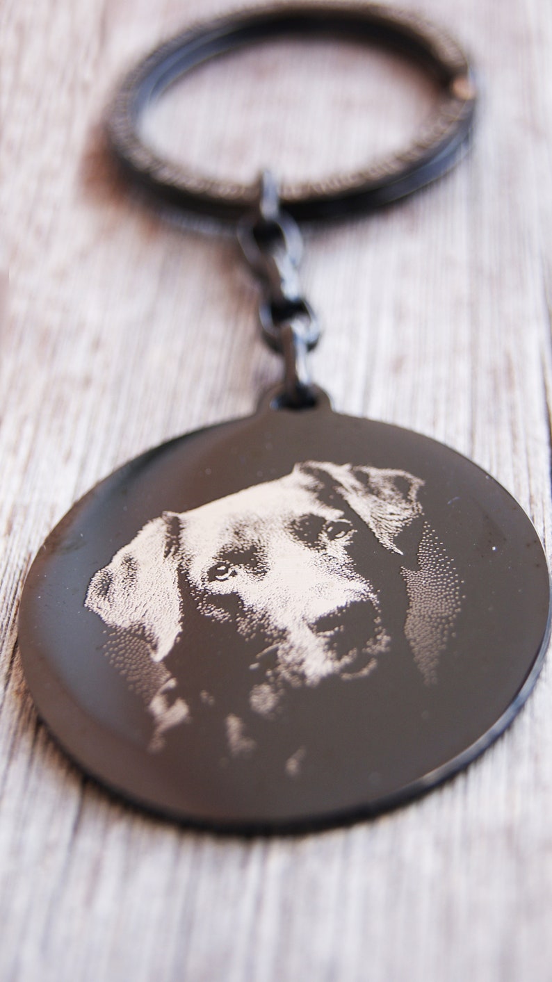 Pode incluir: Um porta-chaves de metal preto com um pendente circular com uma imagem gravada do rosto de um c&atilde;o. O porta-chaves tem um anel e um elo de corrente. O rosto do c&atilde;o &eacute; detalhado com grava&ccedil;&atilde;o clara.