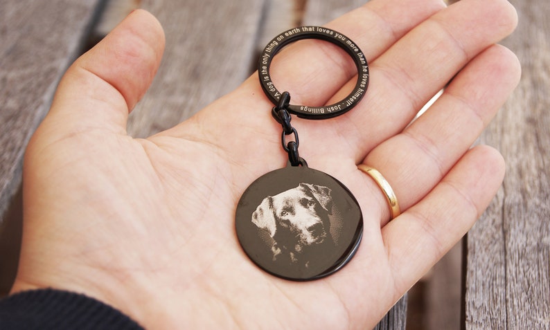 Pode incluir: Porta-chaves de metal preto com um pendente circular com um retrato de c&atilde;o. O anel do porta-chaves tem o texto gravado: "Um c&atilde;o &eacute; a &uacute;nica coisa na terra que te ama mais do que a si mesmo". Uma corrente liga o anel ao pendente.