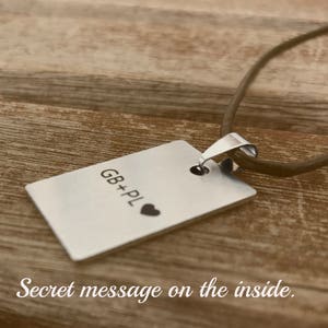 Pu&ograve; includere: Una collana d'argento con un ciondolo rettangolare che dice "GB+PL" con un piccolo simbolo di cuore. La collana &egrave; su una superficie di legno marrone.