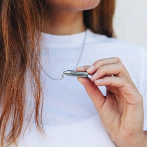 以下が含まれることがあります： ハート型の刻印が施された小さな円筒形のペンダントがついたシルバーネックレス。ペンダントには「Forever in my heart 1956-2022」と刻まれています。