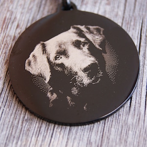 Pode incluir: Uma placa de identifica&ccedil;&atilde;o de c&atilde;o preta e circular com um retrato gravado do rosto de um c&atilde;o. O pelo do c&atilde;o &eacute; de cor clara, contrastando com o fundo escuro. A placa &eacute; provavelmente feita de metal.