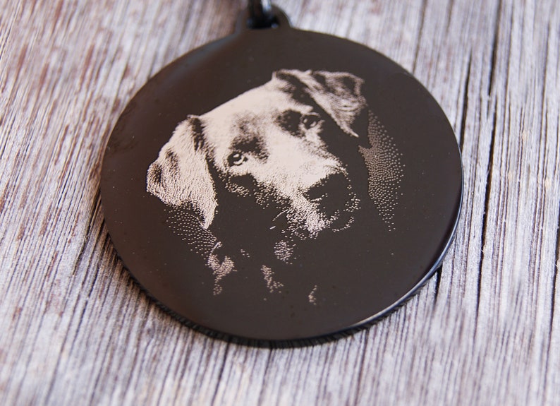 Pode incluir: Uma placa de identifica&ccedil;&atilde;o de c&atilde;o preta e circular com um retrato gravado do rosto de um c&atilde;o. O pelo do c&atilde;o &eacute; de cor clara, contrastando com o fundo escuro. A placa &eacute; provavelmente feita de metal.