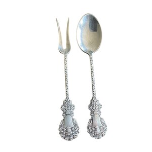 May include: A silver fork and spoon set. The fork has two tines, and the spoon has a rounded bowl. Both utensils have ornate handles with a floral design and a decorative base. The set is likely vintage or antique.