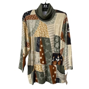 May include: A long-sleeved patchwork tunic with a high, olive green turtleneck. The tunic features a variety of earth-toned fabrics in a patchwork design, including beige, brown, and green. Two pockets are visible on the front. The tunic is hanging on a black hanger.