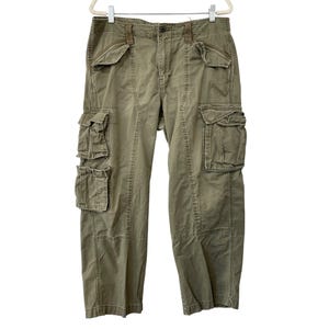 May include: Olive green cargo trousers with multiple pockets on the legs and sides. The trousers are hanging on a white hanger. The trousers have a button and zip closure.
