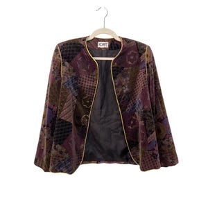 May include: A vintage Koret jacket with a patchwork design in shades of brown, purple, and blue. The jacket has a round collar and is trimmed with gold piping. The interior is lined with a dark fabric. The jacket is hanging on a wooden hanger.