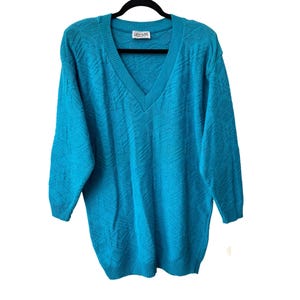 May include: A turquoise V-neck sweater with a textured pattern. The long-sleeved sweater is hanging on a black hanger. The label reads "CARYL BAKER".