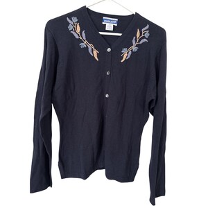 May include: A navy blue cardigan sweater with a V-neck and long sleeves. The sweater features floral embroidery in shades of blue, tan, and gold near the neckline. It has a button-up front and is hanging on a white hanger.