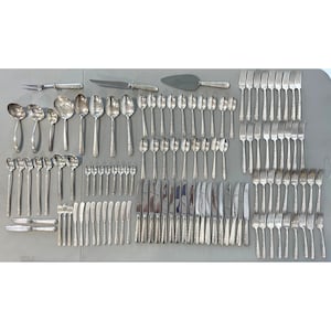 May include: A complete set of silver-colored flatware, including forks, knives, spoons, and serving utensils. The silverware is arranged on a gray surface, showcasing a variety of pieces for a formal dining setting.