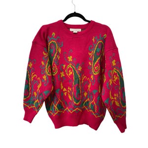 May include: A bright pink sweater with a crew neck and long sleeves. The sweater features a paisley pattern in green and yellow. The sweater is hanging on a black hanger against a white background.