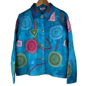 May include: A turquoise button-down jacket with colorful geometric applique designs. The jacket features circles, triangles, and squares in shades of pink, green, and purple, connected by red and blue lines.