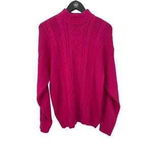 Könnte beinhalten: Ein leuchtend pinkfarbener Pullover mit Rollkragen und langen Ärmeln. Der Pullover hat ein Zopfmuster mit Rautenmustern in der Mitte und strukturiertem Strick an den Seiten. Er hängt an einem schwarzen Kleiderbügel.