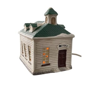 May include: A ceramic schoolhouse figurine with a white exterior and a green roof. The word "SCHOOL" is above the brown door. The windows glow with an orange light, suggesting an interior light. A decorative item.
