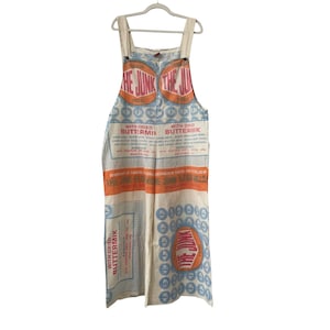 May include: Cream-colored overalls with a unique print featuring the text "THE JUNK FEED" in orange and pink. The design includes blue and orange circular patterns and text blocks, creating a vintage, rustic aesthetic. The overalls hang on a white hanger.