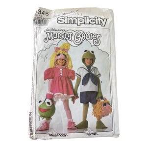 May include: Vintage Simplicity sewing pattern featuring Jim Henson's Muppet Babies. The pattern includes instructions for Miss Piggy and Kermit costumes. The package is white with text and illustrations of the costumes.